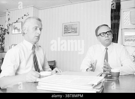 Dir. Public Housing + Councilor De Witte Persons, Whizgle News, Dutch Desk, The Netherlands, 1950 - 2000 on 10-08-1973. The image includes these topics. In a well-lit indoor setting, two men are seated at a table that appears to be made of a light-colored wood. The walls are adorned with vertical stripes of soft shades, creating a subtle backdrop for the scene. One man, with slicked-back silver hair and dressed in a white shirt and a dark tie, looks intently towards the other. He holds a small cup in one hand, possibly a coffee cup. The second man, who sports thick black glasses and a white dr Stock Photo