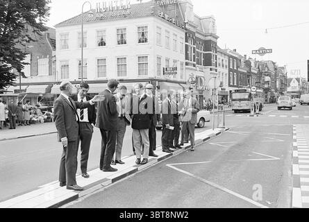Englishmen's walk through the city North South Hollandse Vervoersmaatschappij N.V. NZ, Whizgle News, Dutch Desk, The Netherlands, 1950 - 2000 on 14-07-1969. The image contains these topics. A bustling urban scene unfolds on a busy street lined with two to three-story buildings that display a mix of architectural styles, featuring large windows and ornate facades. The colors of the structures are predominantly muted with soft pastel hues, though some show signs of wear, giving a sense of history. On one side of the street, a diverse group of people stands on the curb, involved in animated discu Stock Photo
