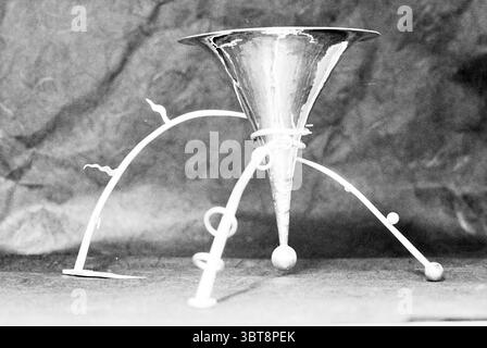 Gedelsmederij Agusto Heemstede Heemstede The Netherlands, Whizgle News, Dutch Desk, The Netherlands, 1950 - 2000 on 06-11-1995. These are the topics in the image. The scene features a striking metallic object that resembles a funnel, prominently placed against a textured backdrop. The funnel has a shiny, reflective surface that catches the light, creating highlights and deep shadows. Its conical shape is complemented by several whimsical, curvilinear legs that seem to be crafted from a lighter material, possibly white or pale in color, adding a playful touch to the composition. The legs extend Stock Photo