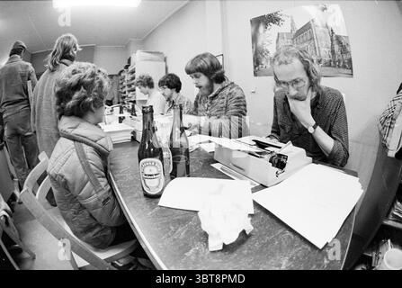 Making Sinterklaas poems Haarlems Dagblad St. Nicholas Sinterklaas, Whizgle News, Dutch Desk, The Netherlands, 1950 - 2000 on 24-11-1978. These are the elements in the image. The scene captures a bustling workspace filled with a diverse group of individuals engaged in various tasks. At the forefront, a woman sits at a wooden table, her back to the viewer, wearing a quilted jacket. Her hair is curly and appears voluminous, reflecting the informal atmosphere. Three men are seated around the table, deeply focused on their activities. One has long hair and a beard, dressed in a patterned sweater. Stock Photo