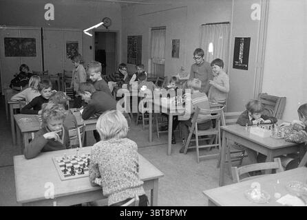 Chess tournament Red Cross IJmuiden Chess, Whizgle News, Dutch Desk, The Netherlands, 1950 - 2000 on 11-04-1972. These are the elements in the image. The scene depicts a lively classroom filled with children engaged in various activities. The room is spacious, with several wooden tables arranged in a somewhat organized manner, each occupied by groups of students. Children are scattered throughout the space, with some sitting at tables focusing intently on games like chess, while others appear to be collaborating on different tasks. The furniture is simple, consisting of sturdy wooden chairs an Stock Photo