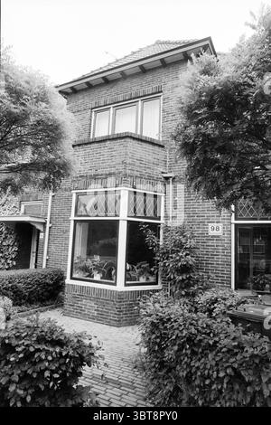 Exterior Herenweg 98 Heemstede Heemstede Herenweg The Netherlands, Whizgle News, Dutch Desk, The Netherlands, 1950 - 2000 on 17-05-1993. These topics appear in the image. The scene presents a charming, two-story brick house characterized by its classic, rectangular design. The facade showcases a rich, deep red-brown color, with a textured pattern that highlights the craftsmanship of the brick. Large windows dominate the front, each framed elegantly and featuring diamond-patterned panes that add a touch of traditional style. Flanking the windows, lush green foliage bursts forth from neatly trim Stock Photo