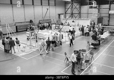 New construction system for houses New construction, Whizgle News, Dutch Desk, The Netherlands, 1950 - 2000 on 13-06-1985. These topics are shown in the image. The scene takes place in a spacious indoor gymnasium, characterized by high ceilings and large windows that allow natural light to stream in. The floor is marked with painted lines and is a polished wood, reflecting the warm tones of the light. In the center, a large construction setup is visible, featuring a partially built structure with several workers actively engaged. They are dressed in a mix of casual and slightly formal attire, Stock Photo