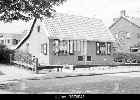 Ext. Klein Beeckesteijn Hoofdstraat Santpoort Santpoort Buildings Santpoort-Noord Hoofdstraat, Whizgle News, Dutch Desk, The Netherlands, 1950 - 2000 on 18-05-1981. These topics are shown in the image. The scene features a charming, modest house with a slanted roof covered in light-colored tiles. The exterior walls are a warm reddish-brown brick, which contrasts nicely with the white shutters that frame the windows, lending a touch of symmetry to the facade. Each window has a unique character, with distinct shapes that enhance the house's overall appeal. In front of the house, a low wooden fen Stock Photo