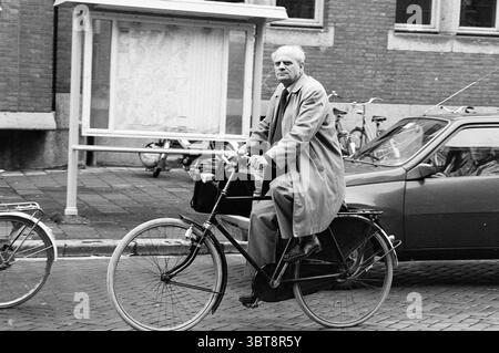 Man on a bicycle in the center of Haarlem., Whizgle News, Dutch Desk, The Netherlands, 1950 - 2000. The image contains these topics. The scene captures a man riding a bicycle in a bustling urban environment. He appears mature, dressed in a long, light-colored coat that billows slightly as he pedals. His hair is gray, adding a touch of wisdom to his demeanor. The bicycle is simple yet well-used, featuring a front basket, and the man sits upright, facing the viewer with a determined expression. Surrounding him, the street is lined with several parked bicycles, indicating a cycling-friendly cultu Stock Photo