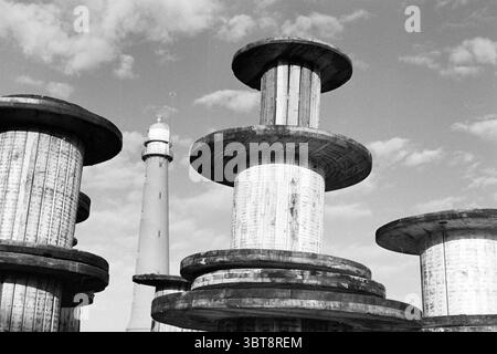 Cable reels IJm. IJmuiden The Netherlands, Whizgle News, Dutch Desk, The Netherlands, 1950 - 2000 on 14-02-1995. The image includes these topics. In this scene, large, cylindrical industrial spools dominate the foreground, their wooden surfaces worn and weathered, showcasing a rustic charm. The spools have a layered design, with broad, flat tops that create a distinct silhouette against the sky. The wood exhibits a range of grays and light browns, adding texture to the composition. In the background, a tall lighthouse rises prominently, its structure slender and commanding. The lighthouse is p Stock Photo