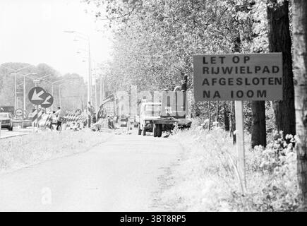 Cycle path work Amsterdamseweg IJmuiden Cycling cycle paths cyclists Work IJmuiden Amsterdamseweg The Netherlands, Whizgle News, Dutch Desk, The Netherlands, 1950 - 2000 on 14-05-1980. These topics appear in the image. The scene features a road alongside a busy construction area. The asphalt appears worn and muted, leading into the distance where it curves. On either side, trees with lush green foliage create a natural boundary, their leaves dappled with sunlight filtering through them, casting shadows onto the road. A circular traffic sign stands prominently, guiding vehicles carefully around Stock Photo