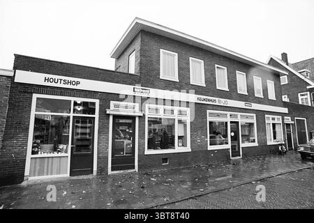 Exterior of Ri-Jo kitchen house., Whizgle News, Dutch Desk, The Netherlands, 1950 - 2000. These topics are shown in the image. The scene features a two-story brick building, characterized by its simple yet functional architecture. The facade is dominated by a warm reddish-brown color, emanating a solid and traditional feel. Several large windows punctuate the front, with some adorned by white trim, allowing glimpses of the interior. The ground floor hosts a series of small storefronts, each with distinct elements that suggest various businesses. The atmosphere feels subdued and slightly melanc Stock Photo