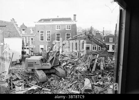 Demolition Winter Garden Demolition demolition demolition yards, Whizgle News, Dutch Desk, The Netherlands, 1950 - 2000 on 24-01-1978. These are the elements in the image. The scene captures a construction area in the midst of demolition. A large excavator, painted in a muted gray, dominates the foreground, its mechanical arm extended as it grips a pile of rubble. The rubble consists of broken bricks, wooden beams, and concrete debris, creating a chaotic yet structured mound indicative of recent destruction. In the background, several buildings stand, their facades showcasing a mix of brick an Stock Photo