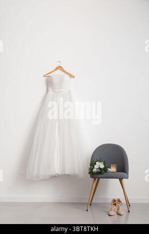 Beautiful wedding dress on white wall, wedding shoes on floor and bridal bouquet on grey chair in room Stock Photo