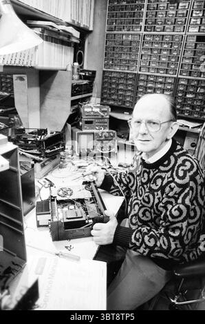 Man in a workshop repairs Philips recorder, Whizgle News, Dutch Desk, The Netherlands, 1950 - 2000 on 05-01-1990. These topics appear in the image. In a cluttered workspace, an older individual is seated at a desk, surrounded by various tools and electronic components. The atmosphere is filled with a sense of industriousness and nostalgia. The person has a thoughtful expression, wearing large, round glasses that reflect the ambient lighting. A patterned sweater, characterized by swirling designs in dark and light tones, adds warmth to the scene, contrasting with the metallic tools scattered on Stock Photo