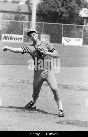 Players Dutch baseball team action Baseball, Whizgle News, Dutch Desk, The Netherlands, 1950 - 2000 on 12-07-1983. The image shows these topics. In the scene, a lone athlete stands on a sun-drenched baseball diamond, captured mid-action as he prepares to pitch a ball. He wears a vibrant uniform in shades of orange and white, adorned with stripes on the sleeves, which emphasizes a classic sporty aesthetic. The fabric looks slightly worn, suggesting he may be a seasoned player. His posture is dynamic and focused; his throwing arm is drawn back, indicating a moment of concentration before the thr Stock Photo