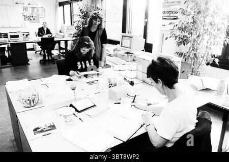 Haarlems Dagblad layout and promotion Haarlem The Netherlands, Whizgle News, Dutch Desk, The Netherlands, 1950 - 2000 on 29-06-1998. The image shows these topics. The scene depicts an office environment characterized by a blend of individuals engaged in various tasks. In the foreground, two women sit at a large, cluttered desk. One, with long, dark hair, appears focused on writing or reviewing documents, while the other, with short hair, is deeply engrossed in her work, taking notes or filling out forms, suggesting a collaborative effort. Both are surrounded by stacks of papers and office supp Stock Photo