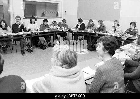 Class of school Social Labor Schools, Whizgle News, Dutch Desk, The Netherlands, 1950 - 2000 on 17-12-1981. These are the elements in the image. In a classroom setting, a group of students is seated at long, rectangular tables arranged in a slightly curved formation, suggesting a collaborative atmosphere. The room appears well-lit, with soft, ambient light illuminating the space, casting gentle shadows across the surfaces. On one side, a cluster of individuals sits closely together, engaged in discussion. Their expressions range from concentration to curiosity, indicating a dynamic exchange of Stock Photo