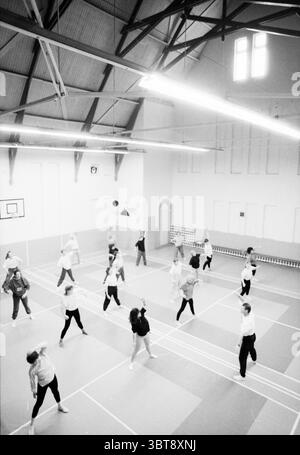 Gym in Koek Haarlem Haarlem The Netherlands, Whizgle News, Dutch Desk, The Netherlands, 1950 - 2000 on 08-01-1991. These topics appear in the image. The scene captures a spacious gymnasium characterized by its high ceilings and exposed wooden beams. Natural light streams in from tall windows, casting soft shadows across the polished wooden floor. The color palette is predominantly muted, with shades of gray and white dominating the walls, juxtaposed with the dark wooden beams and the lighter wooden flooring. A group of dancers fills the space, their movements dynamic and expressive. They are d Stock Photo