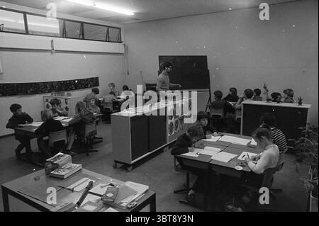 4 school classes in a classroom IJmuiden Schools, Whizgle News, Dutch Desk, The Netherlands, 1950 - 2000 on 11-01-1972. These are the elements in the image. The scene captures a school classroom filled with activity and engagement. The room is spacious and well-lit, with natural light filtering through large windows positioned near the ceiling. The walls are adorned with various educational posters and displays, hinting at a creative learning environment. In the foreground, several groups of children are seated at small, wooden desks, each covered with sheets of paper and school supplies. The Stock Photo