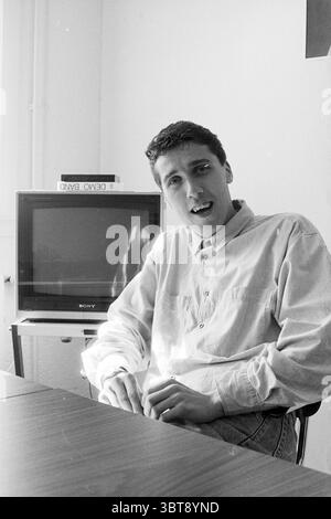 Portrait radio DJ, Whizgle News, Dutch Desk, The Netherlands, 1950 - 2000 on 03-1990. These are the topics in the image. In a bright, minimalist room, a young man sits casually in a chair, leaning slightly forward with a relaxed yet engaged expression. He has short, dark hair styled with a soft wave and is dressed in a light-colored, button-up shirt with the sleeves rolled up to the elbows, suggesting a casual atmosphere. Behind him, a small television set sits on a narrow, wheeled table, its screen reflecting the light from the window, while an item labeled “DEMO BAND” rests on top. The walls Stock Photo