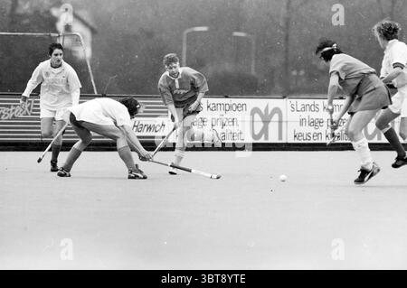 Bloemendaal-Wageningen Hockey Wageningen, Whizgle News, Dutch Desk, The Netherlands, 1950 - 2000 on 13-03-1988. The image contains these topics. The scene captures a dynamic moment on a field, where a group of players is engaged in a hockey match. The landscape is marked by a muted, monochromatic palette dominated by shades of gray, emphasizing the intensity of the competition. In the foreground, two players are locked in a struggle for control of the ball. One is crouched down, stick poised to intercept, while the other stands tall, looking determined. The crouching player wears a dark unifor Stock Photo