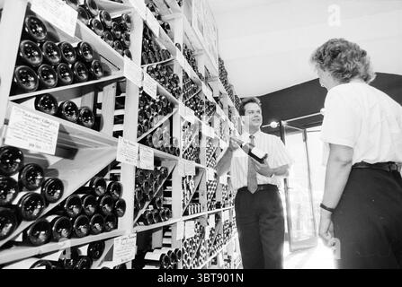 Wine shop Bacchus, Whizgle News, Dutch Desk, The Netherlands, 1950 - 2000 on 03-09-1991. The image includes these topics. In a spacious, well-lit room, a man and a woman engage in conversation amidst an impressive display of wine bottles. The wall behind them is adorned with neatly arranged wooden racks, showcasing a variety of dark glass bottles, their labels not visible. The man, wearing a light-colored striped shirt and dark pants, holds a bottle in one hand while gesturing with the other, suggesting a sense of enthusiasm and engagement. The woman, dressed in a casual white blouse and dark Stock Photo