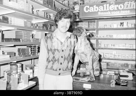 Mark de Groot in store Shops and kiosks, Whizgle News, Dutch Desk, The Netherlands, 1950 - 2000 on 24-02-1983. The image includes these topics. In a cozy shop filled with shelves stocked with various items, a man stands confidently next to a dog, both positioned at the center of the scene. The man has a distinctive mustache and wears a patterned vest adorned with rich colors like deep green, brown, and hints of orange. His hair is curly and modestly styled, giving him a classic, nostalgic appearance. The dog, a medium-sized breed with a sleek coat in shades of brown and black, sits comfortably Stock Photo