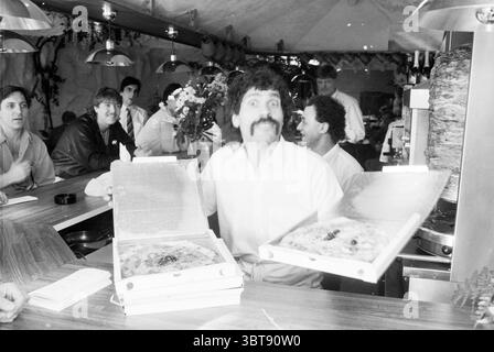 Italian restaurant Nieuw Vennep Restaurant Nieuw-Vennep, Whizgle News, Dutch Desk, The Netherlands, 1950 - 2000 on 02-04-1988. The image contains these topics. The scene captures a lively restaurant setting filled with a mix of people and vibrant culinary activity. The central figure, a man with a prominent mustache, is animatedly presenting two pizza boxes, one in each hand. He wears a light-colored shirt that contrasts with the darker tones of the wood-paneled countertop. The background features a group of patrons seated at wooden tables, enjoying their meals and engaging in conversation. Th Stock Photo
