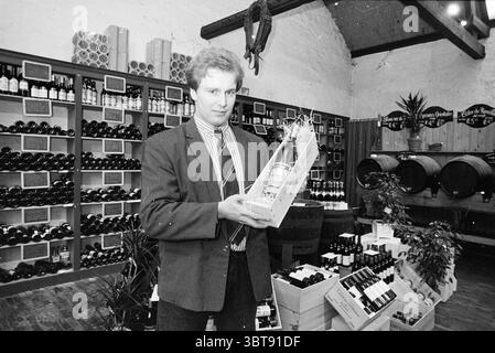 Wine shop Molenaar Bloemendaal Wine Bloemendaal Zomerzorgerlaan, Whizgle News, Dutch Desk, The Netherlands, 1950 - 2000 on 19-01-1989. These are the elements in the image. The scene captures a retail space dedicated to wine, characterized by rustic charm and a cozy atmosphere. In the foreground, a man stands holding a wrapped bottle, exuding a sense of pride or excitement. He is dressed in a sharp suit with a striped tie, suggesting a blend of professionalism and casual observance of the setting. Surrounding him, shelves are densely packed with various bottles of wine, their dark glass glisten Stock Photo