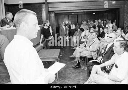 Mr. Dam at auction Restaurant Auction, Whizgle News, Dutch Desk, The Netherlands, 1950 - 2000 on 07-07-1970. These topics are shown in the image. In a spacious indoor setting, a crowd is gathered, with a mix of well-dressed individuals and more casually attired attendees. The room features wooden paneling and an ornate floral-patterned wallpaper that suggests an old-world charm. Natural light streams in from a partially visible window, illuminating the faces of the people, creating a warm yet serious atmosphere. At the forefront, a man stands with his back to the viewer, holding a sheet of pap Stock Photo