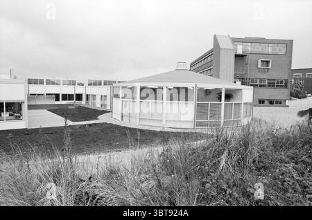 Newly built Heliomare school Wijk aan Zee Wijk aan Zee, Whizgle News, Dutch Desk, The Netherlands, 1950 - 2000 on 22-09-1991. These topics appear in the image. The scene presents a modern architectural landscape characterized by a mix of angular and circular structures. In the foreground, a round building with a sloped, transparent roof stands out, its glass panels reflecting the cloudy sky above. Surrounding it, patches of green grass and low shrubs create a natural contrast against the stark white of the building. To the right, a taller, rectangular structure looms, featuring a combination o Stock Photo