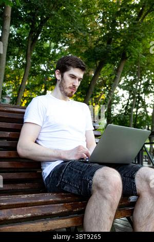 Casual man sitting in park with laptop. Typing and working remotely outside. concept of remote work, flexibility, and digital freedom in a nature Stock Photo