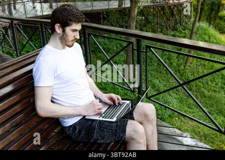 Freelancer using computer outdoors in city park. White t-shirt, black shorts, relaxed setting. Stock Photo