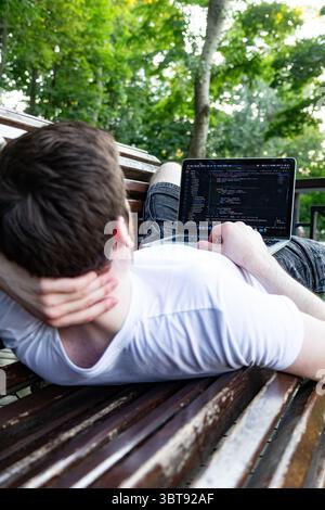 Man enjoying remote work in park. Laptop on lap, wearing white t-shirt, relaxing outdoors. Stock Photo