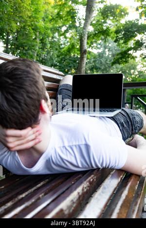 Focus on laptop screen with empty space for photo or content, while a young man in casual outfit works on computer in park. Green trees Stock Photo