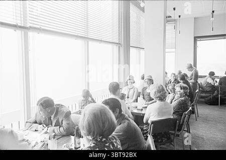 People look at view from Bouwes Palace Zandvoort, Whizgle News, Dutch Desk, The Netherlands, 1950 - 2000 on 22-08-1965. These topics are shown in the image. The scene captures a lively dining area filled with groups of people engaged in conversation. Large windows stretch along one side, allowing soft, diffused natural light to fill the space, creating a bright yet cozy atmosphere. The tables are set with simple white tablecloths, and some people are dining while others appear to be deep in discussion. The room is adorned with contemporary decor, featuring minimalistic furnishings. A mix of pa Stock Photo