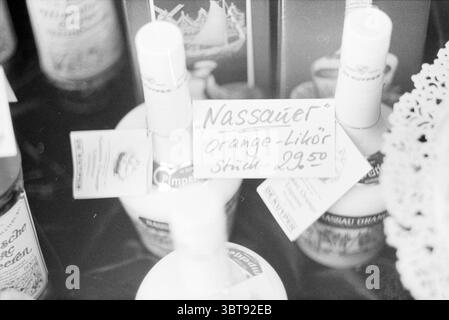 Photo impression of the town of Nassau in Germany Nassau Germany, Whizgle News, Dutch Desk, The Netherlands, 1950 - 2000 on 09-09-1980. These are the topics in the image. The scene depicts a collection of various bottles sitting closely together, likely on a display surface such as a table or a shelf. The bottles are made of glass with a range of shapes, some rounded and others more angular. Most are clear, allowing the contents to be seen, while a few have colored liquids that impart a subtle hint of their flavors. Among the bottles, there are several with caps that might be cork or metal, le Stock Photo