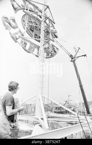 Placing neon advertising at Loe Lap Amsterdam Advertising, Whizgle News, Dutch Desk, The Netherlands, 1950 - 2000 on 25-05-1976. The image contains these topics. In this scene, a man stands in the foreground, dressed in a plain, short-sleeved shirt and focused intently on an intricate structure above him. His posture suggests concentration, with one hand poised as if contemplating or adjusting something in his grasp. Above him, a large, circular framework, possibly a sign or decorative element, dominates the upper part of the view. The structure is composed of metal, displaying a stark white c Stock Photo