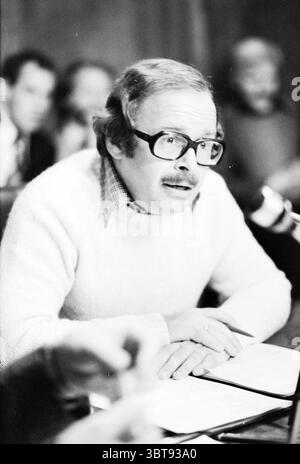 Portrait of councilor Haarlemmerliede and Spaarnwoude in council chamber Haarlemmerliede en Spaarnwoude, Whizgle News, Dutch Desk, The Netherlands, 1950 - 2000 on 17-10-1978. These topics are shown in the image. In the scene, a man with a light-colored sweater sits at a table, speaking intently into a microphone. He has thick glasses and a mustache, his expression reflecting seriousness and focus. The table is adorned with scattered papers, possibly notes or documents he's referencing. Behind him, several figures are visible, although they appear somewhat blurred, suggesting they are part of a Stock Photo