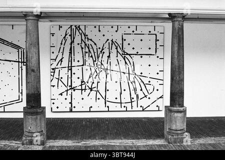 Expo Vishal Noud van Dun, Whizgle News, Dutch Desk, The Netherlands, 1950 - 2000 on 12-01-1995. These are the elements in the image. The scene features a large, abstract artwork prominently displayed on a white wall. The artwork consists of bold, black lines that create a complex network of geometric shapes and patterns, resembling a map or a series of interconnected paths. These lines interweave across the canvas, forming a striking contrast against the bright background. In the foreground, two large, cylindrical columns rise, their surfaces textured and displaying hints of age. These columns Stock Photo