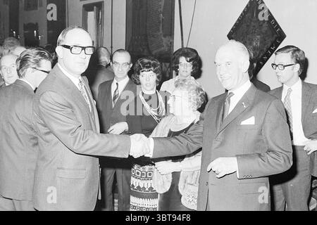 A jubilee is congratulated, Whizgle News, Dutch Desk, The Netherlands, 1950 - 2000 in 1972. The image shows these topics. In a well-lit indoor setting, a group of individuals is gathered, creating an atmosphere of formal social interaction. The focus is on two men in the foreground, both engaged in a handshake, symbolizing a moment of agreement or greeting. One man wears large, dark-framed glasses and a suit, exuding a sense of authority, while the other, dressed in a suit as well, smiles slightly, conveying approachability. Surrounding them, a mixture of men and women contributes to the scene Stock Photo