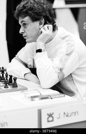 Blastovens Chess Tournament J. Nunn, Whizgle News, Dutch Desk, The Netherlands, 1950 - 2000 on 29-01-1991. These are the elements in the image. The scene captures a focused moment in a chess match, showcasing an individual deeply immersed in thought. The person, a young male with curly hair, is seated at a table, leaning slightly forward with a pensive expression. His elbow rests against the table, while his hand supports his chin, suggesting concentration and contemplation. The table is neatly arranged, featuring a chessboard at its center where wooden pieces are positioned, some clearly move Stock Photo