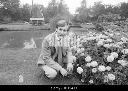Mr Leo Roozen in the garden Vogelenzang Gardens and parks Vogelenzang, Whizgle News, Dutch Desk, The Netherlands, 1950 - 2000 on 31-08-1983. These are the topics in the image. A serene garden scene unfolds, characterized by a lush expanse of greenery meticulously maintained. In the foreground, a man, dressed in a light gray suit with a subtly textured blazer, is kneeling beside a vibrant display of flowers. The blooms are a mix of soft colors—whites and pale yellows—that stand out against the rich green of the foliage and grass. The background reveals a tranquil pond, its surface reflecting th Stock Photo