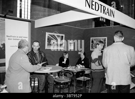Nedtrain stand at the Alkmaar job market Alkmaar, Whizgle News, Dutch Desk, The Netherlands, 1950 - 2000 on 21-11-2001. These are the topics in the image. In a spacious indoor setting, a gathering is taking place that suggests an event focused on transportation or tourism. The area is adorned with a backdrop featuring a prominent booth, which showcases a simple yet bold color scheme of dark green walls and a lighter foreground, giving it a sophisticated appearance. The lighting is soft, creating a warm and inviting atmosphere. Several people are engaged in conversation, standing at small table Stock Photo