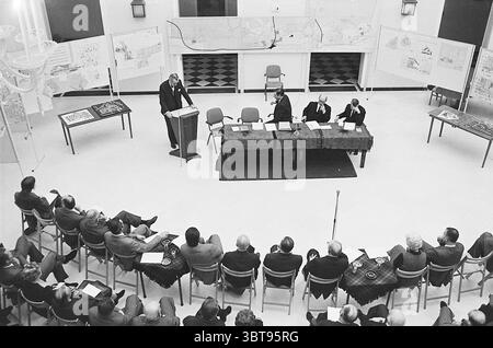 Hearing Bloemendaal Meetings, Whizgle News, Dutch Desk, The Netherlands, 1950 - 2000 on 04-02-1971. These topics are shown in the image. The scene presents a gathering in a spacious, well-lit room with an atmosphere of seriousness and engagement. At the center, a long table covered with a dark cloth is positioned, surrounded by several individuals in formal attire. These figures appear to be deep in conversation or discussion, suggesting a formal meeting or presentation. In the foreground, a speaker stands at a podium, gesturing with one hand and appearing to address the group with authority. Stock Photo
