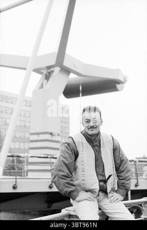 Supervisor Lange Brug Mr. Eskam, Whizgle News, Dutch Desk, The Netherlands, 1950 - 2000 on 18-01-1995. These topics appear in the image. In this scene, a man sits casually on a railing, showcasing a relaxed posture. He has short, dark hair and a mustache, contributing to a confident demeanor. His attire consists of a light-colored shirt layered with a bright orange vest that stands out against the muted backdrop. The setting features industrial elements, particularly a large, abstract structure resembling a crane in the background, painted in neutral tones with accents of red and white. This s Stock Photo