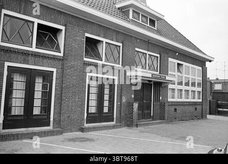 Ext. music school IJmuiden Exterieur, Whizgle News, Dutch Desk, The Netherlands, 1950 - 2000 on 23-05-1972. These topics are shown in the image. The scene presents a brick building characterized by a classic architectural style, featuring a combination of dark and light tones. The structure includes a prominent entrance marked by a rectangular awning that extends outward. Window panes are intricately designed, displaying a diamond pattern that adds visual interest. The building's exterior is primarily a deep brown, accentuated by white window frames and detailing, creating a striking contrast. Stock Photo