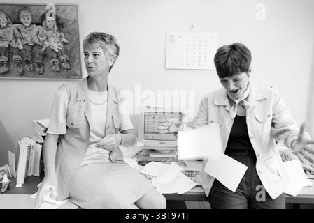 Two ladies A.W.V.N. Haarlem Westelijke Randweg The Netherlands, Whizgle News, Dutch Desk, The Netherlands, 1950 - 2000 on 28-05-1997. These topics appear in the image. In a brightly lit office space, two women are engaged in a lively conversation. To the left, one woman has short, light-colored hair styled neatly and wears a tailored light gray vest over a white blouse. She has an expressive demeanor, with one eyebrow raised and a slight smile, suggesting amusement or disbelief. On the right, the second woman exudes energy, with a slightly shorter, darker bob hairstyle. She is dressed in a lig Stock Photo