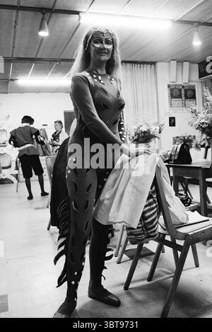 Shakespeare: dressing up actors Dr. Broekema for the public Theater Diever Hezenes, Whizgle News, Dutch Desk, The Netherlands, 1950 - 2000 on 06-08-1977. The image includes these topics. In a vibrant indoor setting, a person stands confidently, dressed in a striking costume characterized by intricate patterns and eye-catching details. The costume has a deep hue, possibly a mix of dark blues or purples, accentuated with silver or metallic embellishments that catch the light and add a dynamic quality to the outfit. The subject's makeup complements their look, with bold colors around the eyes and Stock Photo