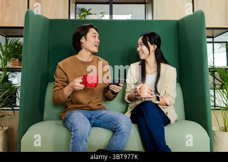 Asian coworkers sitting on green sofa in office lounge holding ceramic mugs and smartphone Stock Photo
