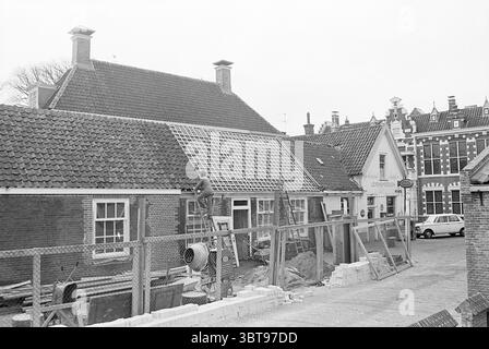 Report Oud Velsen Report, Whizgle News, Dutch Desk, The Netherlands, 1950 - 2000 on 08-03-1968. These topics are shown in the image. The scene depicts a construction site in an urban setting, showcasing a blend of historical and contemporary architecture. On the left, a worker is perched on a ladder, carefully aligning roofing tiles on the steep slanted roof of a traditional brick building. He is dressed in practical work clothes, and a focused expression hints at his concentration on the task. The foreground features a wooden fence, enclosing the work area and adding a sense of separation fro Stock Photo