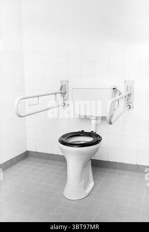 Cross association building Kwadijk Cross associations Kwadijk, Whizgle News, Dutch Desk, The Netherlands, 1950 - 2000 on 22-04-1980. These topics are shown in the image. The scene features a simple, utilitarian restroom setting. Dominating the foreground is a white ceramic toilet bowl with a black lid. It is positioned against a wall that is covered in smooth, white tiles, giving a clean and sterile atmosphere. On either side of the toilet, there are support rails, also white, that extend from the wall to provide assistance. These rails are designed with a minimalist aesthetic, adding to the f Stock Photo