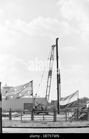 First pile of Phillips building Oudeweg Haarlem. First pile Haarlem Oudeweg The Netherlands, Whizgle News, Dutch Desk, The Netherlands, 1950 - 2000 on 10-09-1974. These are the elements in the image. The scene features a prominent industrial setting, characterized by a large crane positioned centrally, towering above the surrounding elements. The crane's metallic structure is painted in shades of grey and yellow, reflecting its utilitarian purpose. It is actively engaged in lifting a vertical metal beam, which stands upright against the sky. In the foreground, two large flags flutter in the wi Stock Photo
